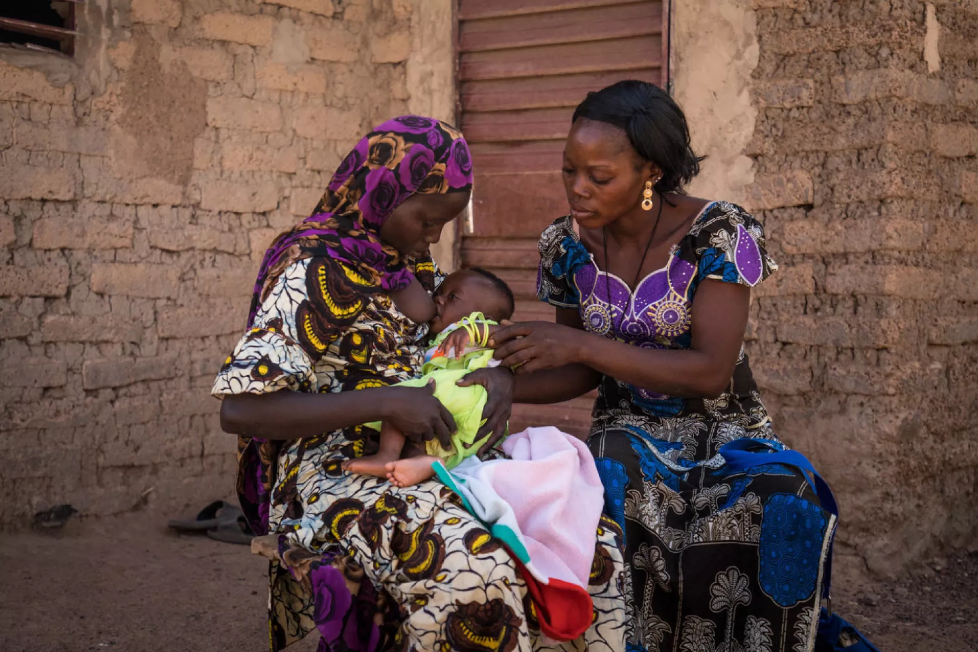 Two women in colourful dresses sitting outside a brick building, one woman breastfeeding a baby
