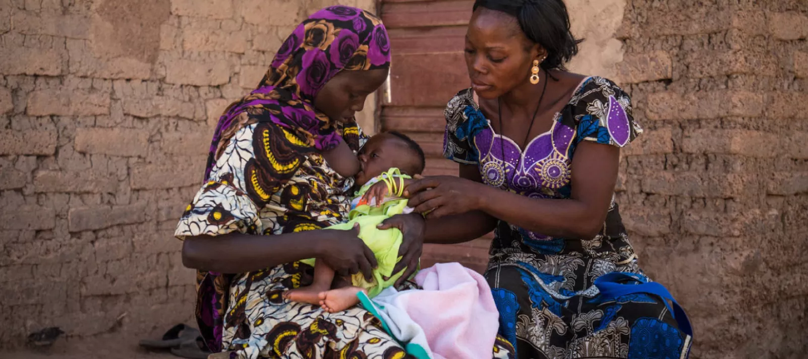 Two women in colourful dresses sitting outside a brick building, one woman breastfeeding a baby